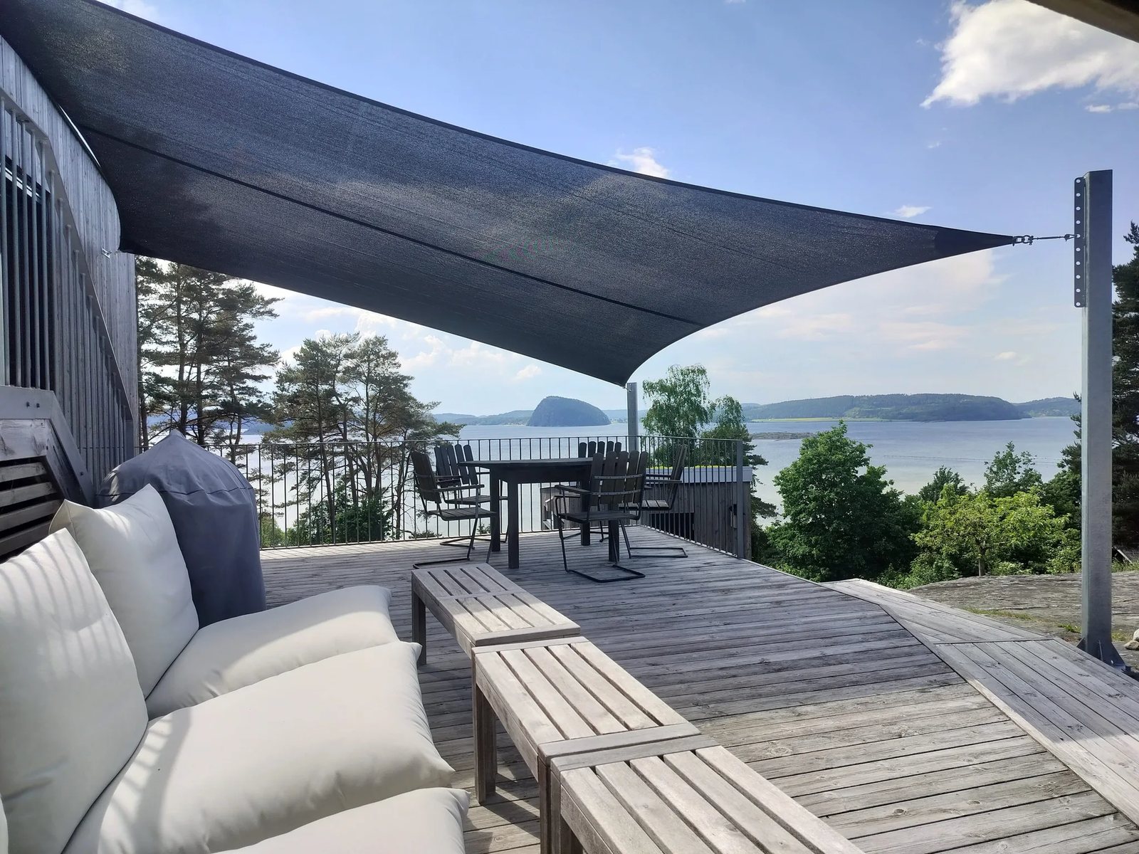 Mörkt grått solsegel spänt över träaltan med loungemöbler och havsutsikt